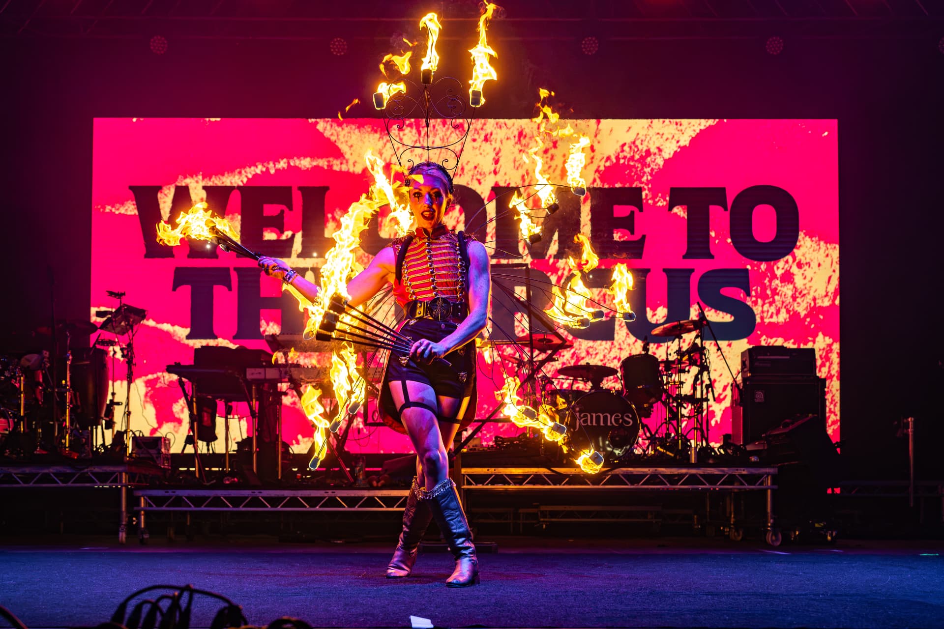 A fire performer in a red ringmaster waistcoat performs in front of a band on stage at Rock N Roll Circus