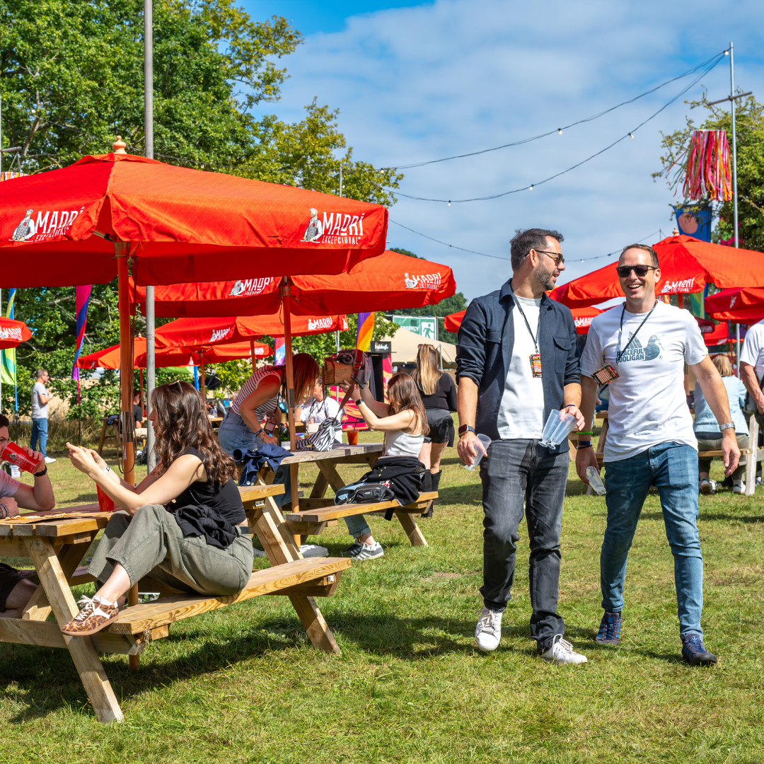 Festivalgoers relaxing around picnic tables on a sunny lawn beneath red parasols
