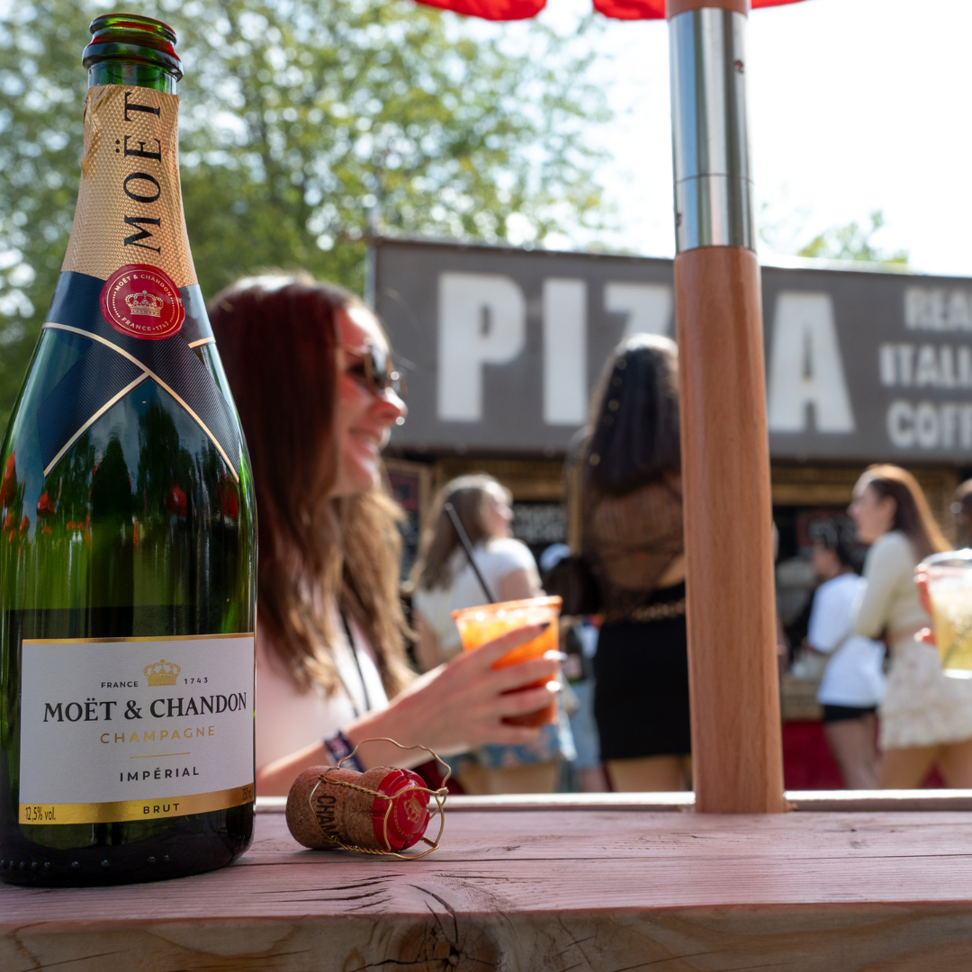 A bottle of Moët & Chandon champagne and its cork on a wooden picnic table, with festivalgoers holding drinks beside a pizza stand