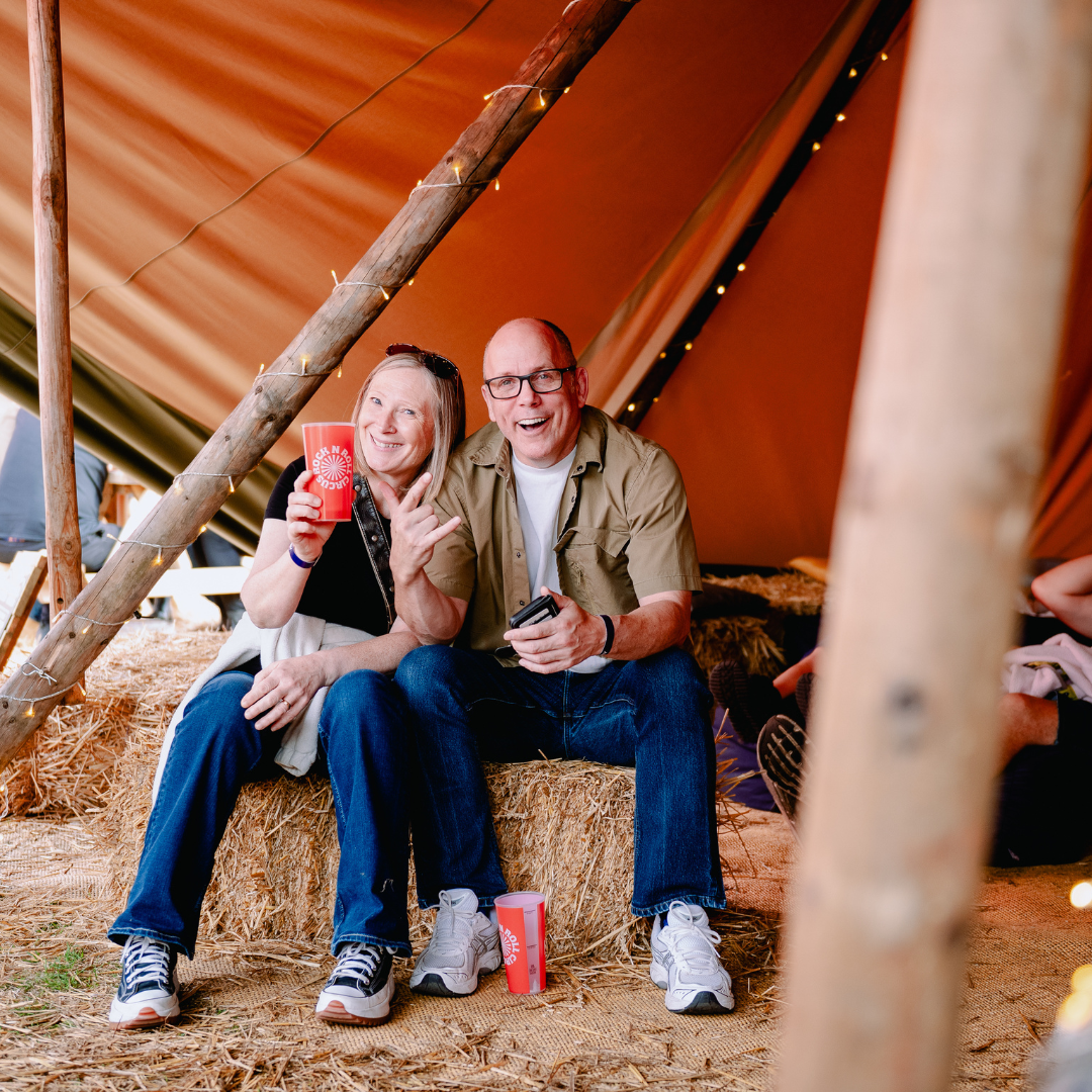 A smiling festivalgoer and companion seated on hay bales inside a fairy-lit tipi, holding Rock N Roll Circus cups