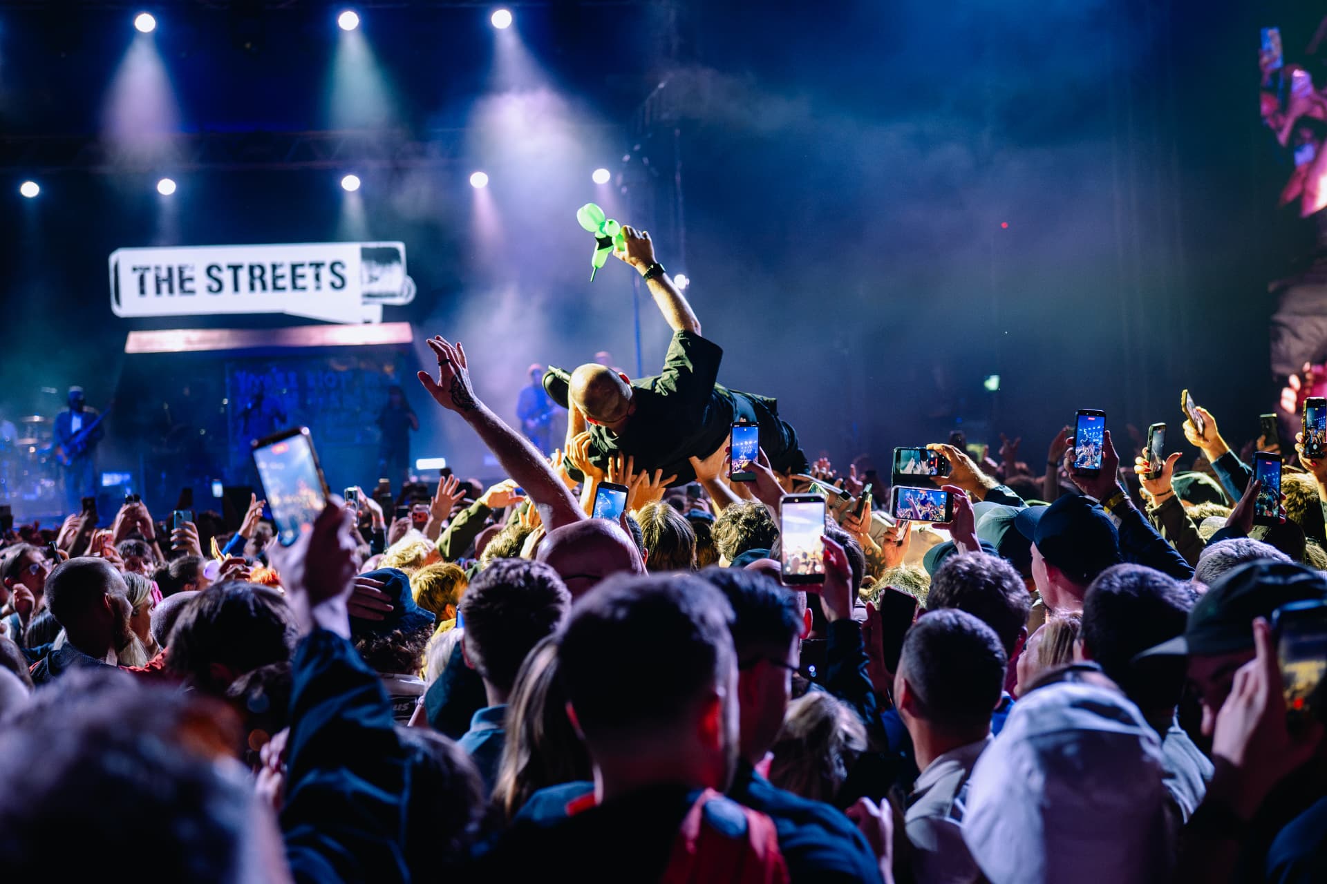 A performer crowd-surfing above raised hands under blue stage lights