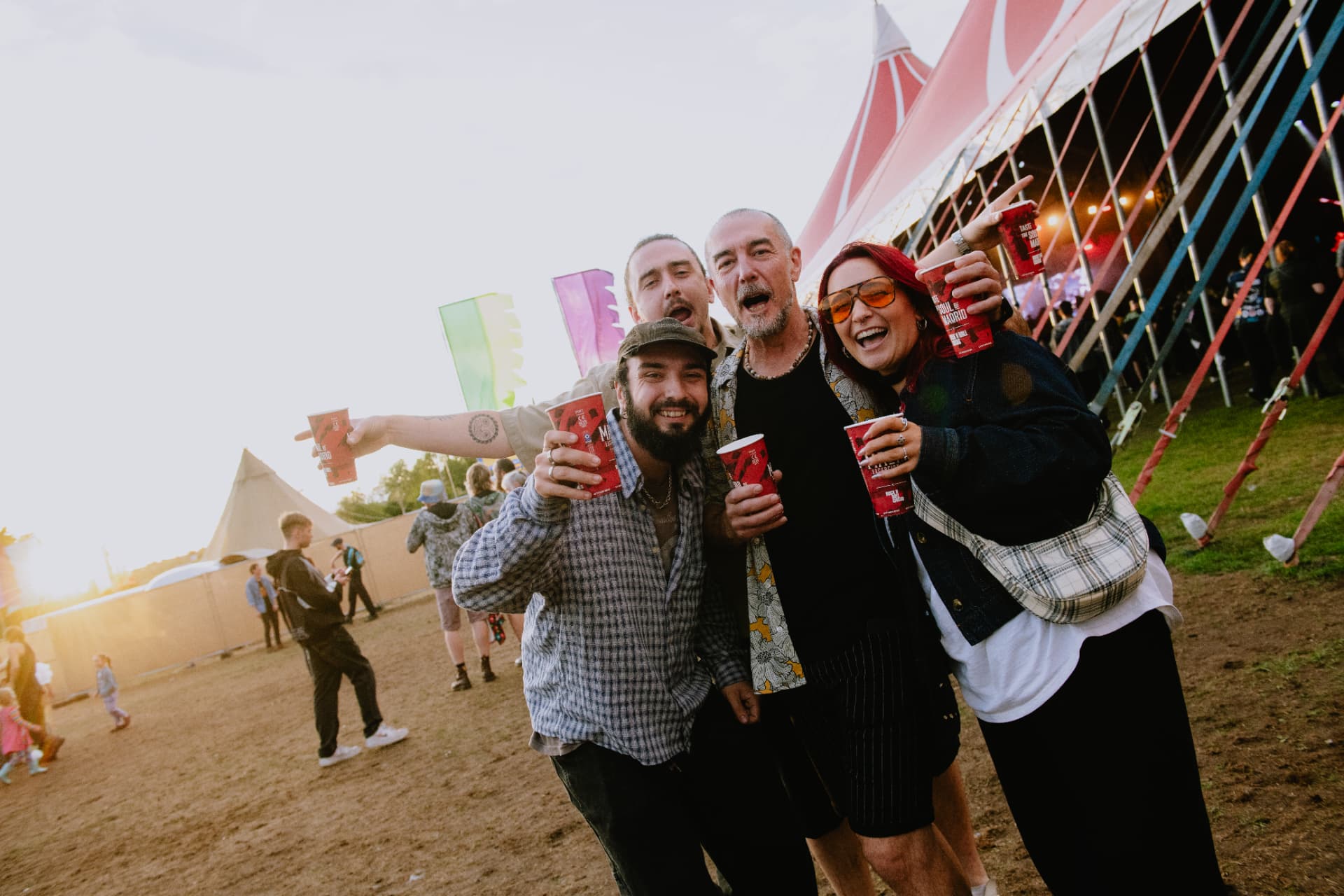 Four festival-goers cheering with drinks in front of the Rock N Roll Circus big top